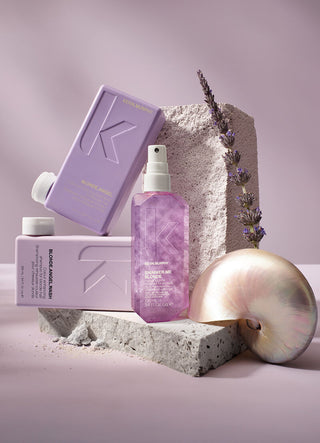 Kevin Murphey product set, the shampoo is in a light pink bottle, conditioner is in a rose pink bottle, and the shimmer spray is in a translucent bottle but the liquid is purple. The products are on top of a rock and there is lavender and a pearl sea shell balancing on rock