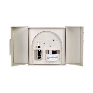 Light yellow/beige box open to showcase products inside. The serum in a gold dropper bottle and the cream in a white jar with gold lid. The box is against a white background