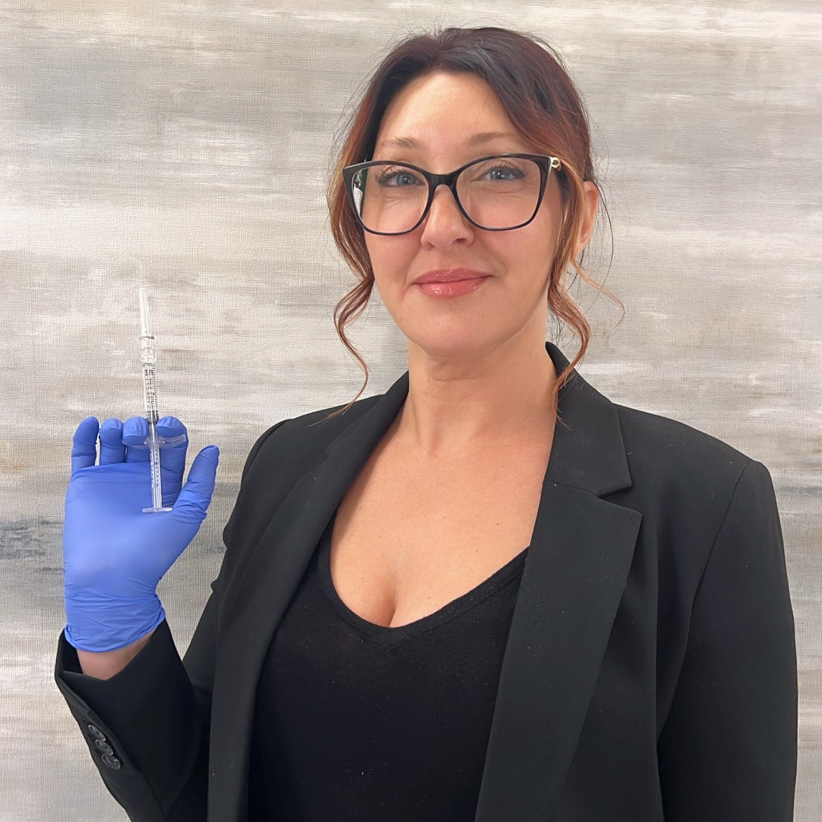 Woman in black blazer holding a syringe against a gray background