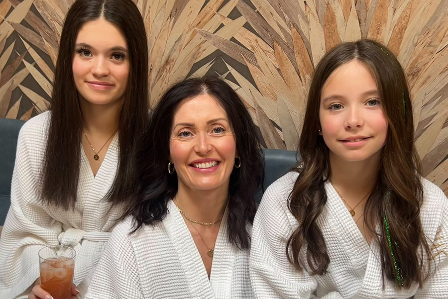 A woman and her 2 daughters in white robes sitting together with a wooden wall background