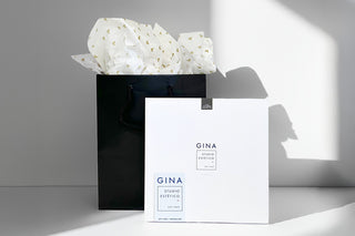 Black gift bag with white tissue paper with small gold hearts and a white spa pamphlet labeled 'GINA' on a gray background.